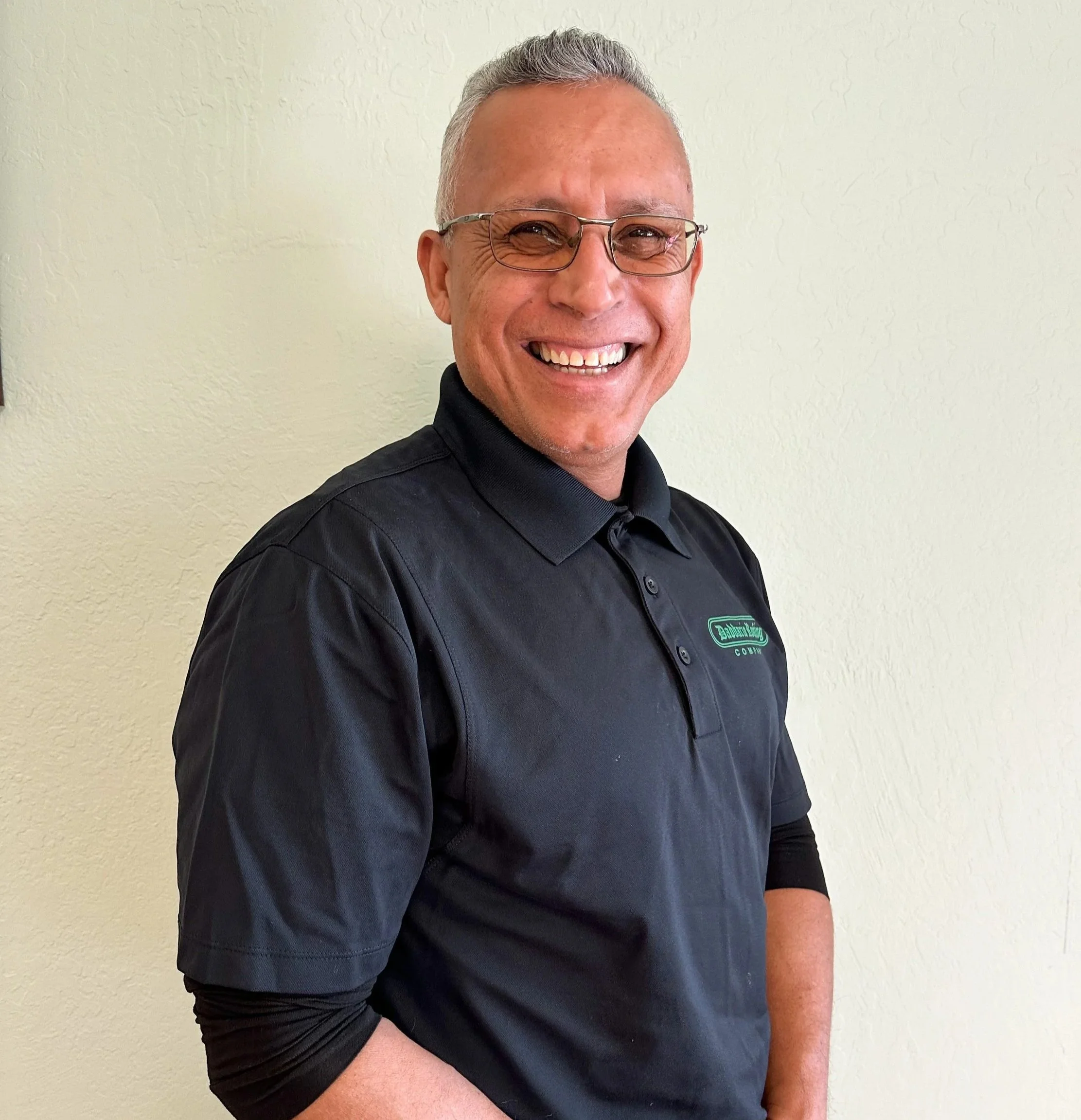 A smiling man with glasses wearing a black collared shirt standing against a plain light-colored wall.
