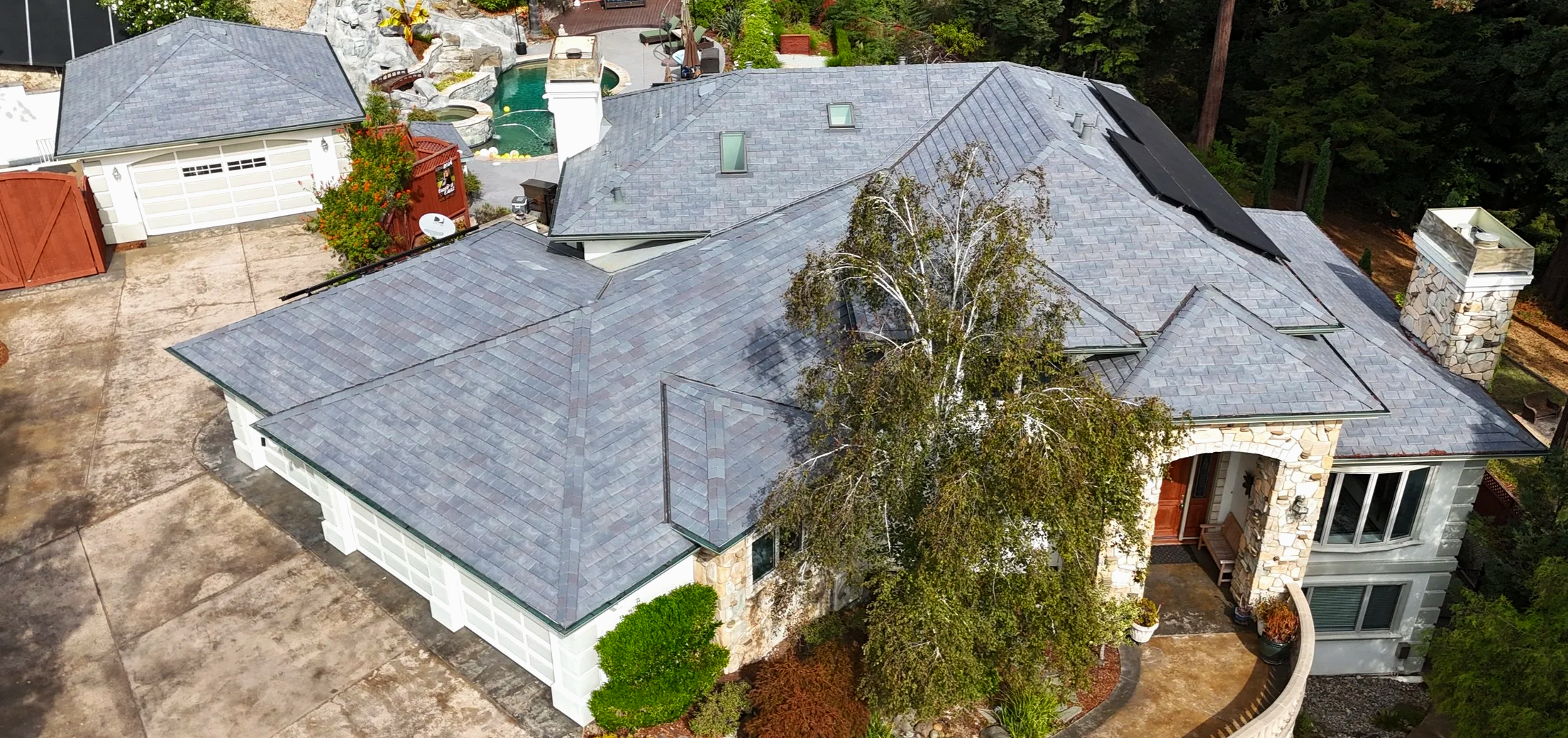 Front view of a house with a brick and green exterior, large windows, a garage door, and a landscaped garden with bushes, small trees, and brick planters.