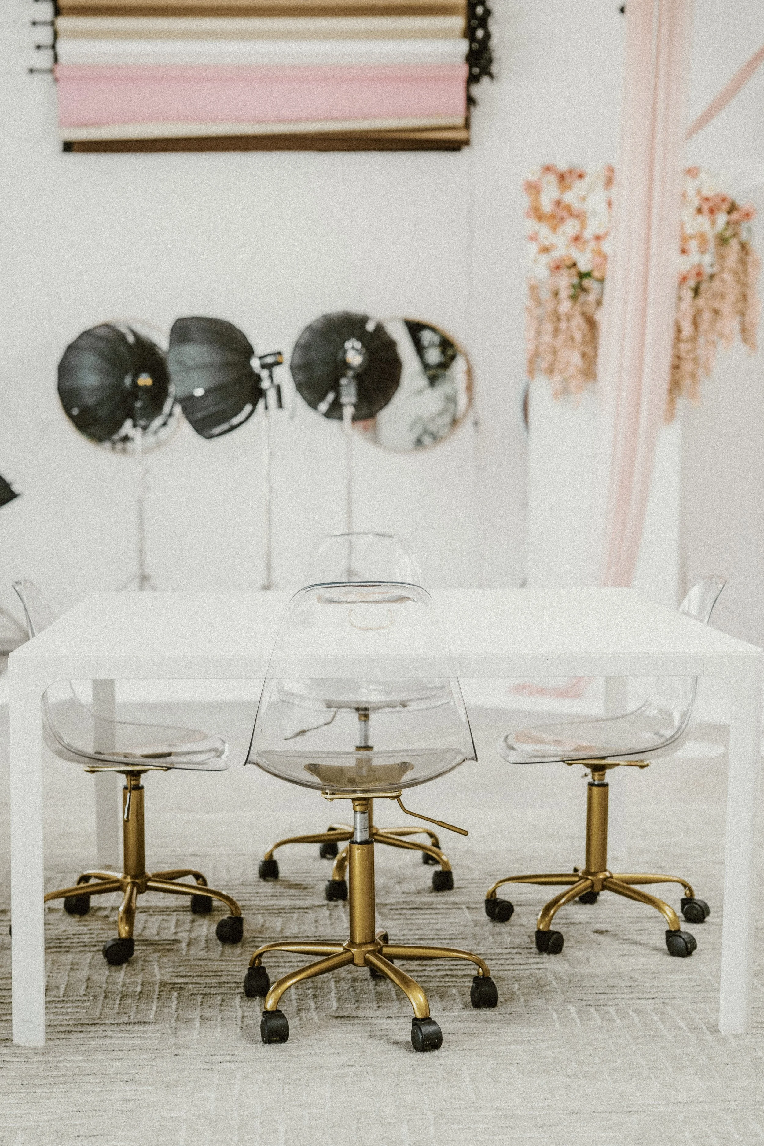 A modern, minimalistic office space with a white table and four white swivel chairs, a vase of pink flowers on the table, and a green sofa with yellow ottomans in the background. There are round mirrors and a neon sign that says 