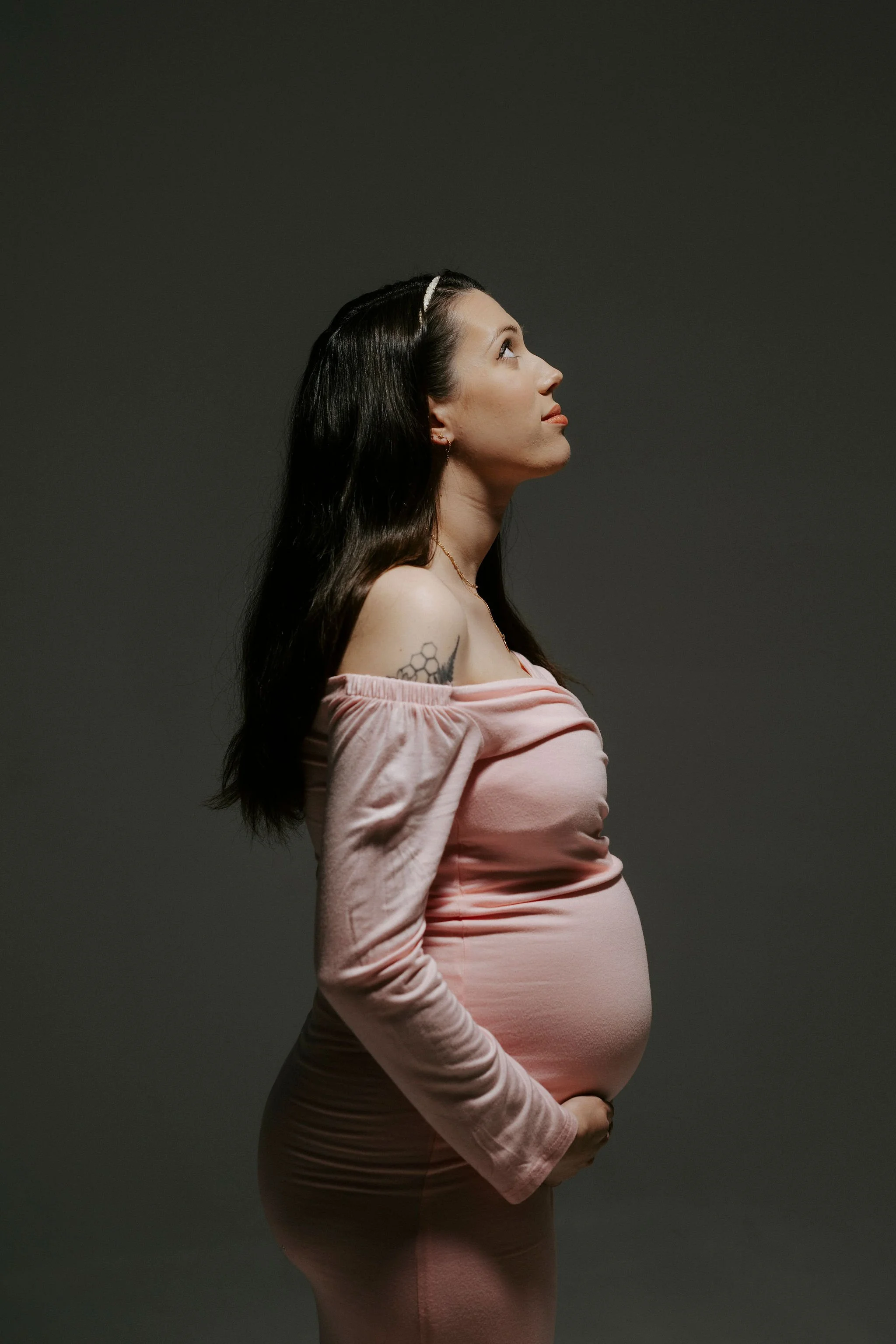 Side profile of a pregnant woman with long dark hair, wearing a pink off-shoulder top and matching pants, standing against a dark background.