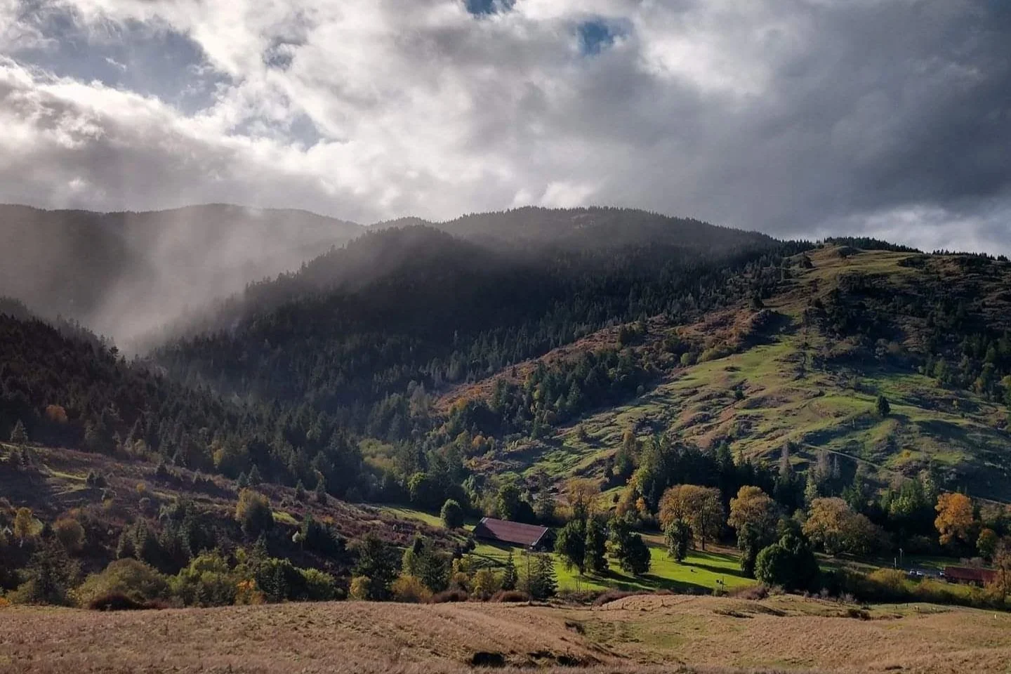 Scenic view of rolling green hills and mountains with trees, clouds, and sunlight breaking through the clouds.
