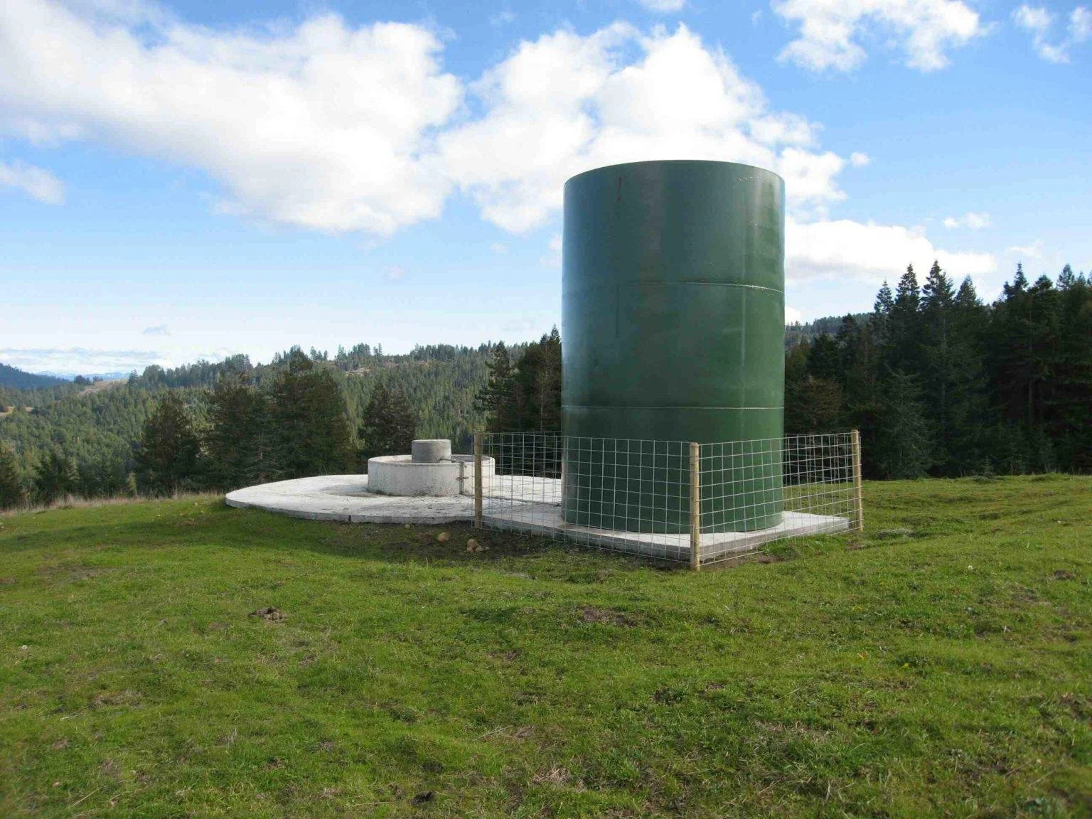 A large green cylindrical structure on a grassy hill with a concrete base, surrounded by a metal fence, with forested hills and a partly cloudy blue sky in the background.