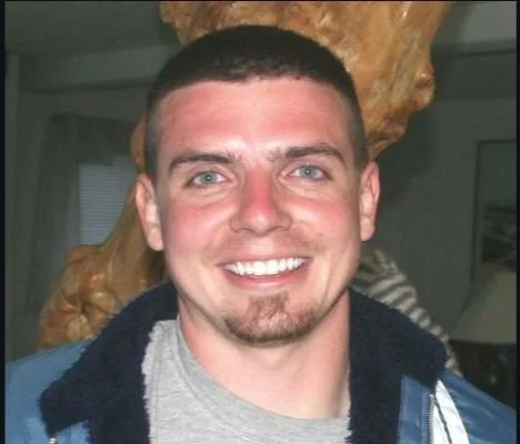 A smiling young man with short dark hair and a goatee, wearing a gray shirt and a dark jacket, indoors with a wooden sculpture or decor in the background.