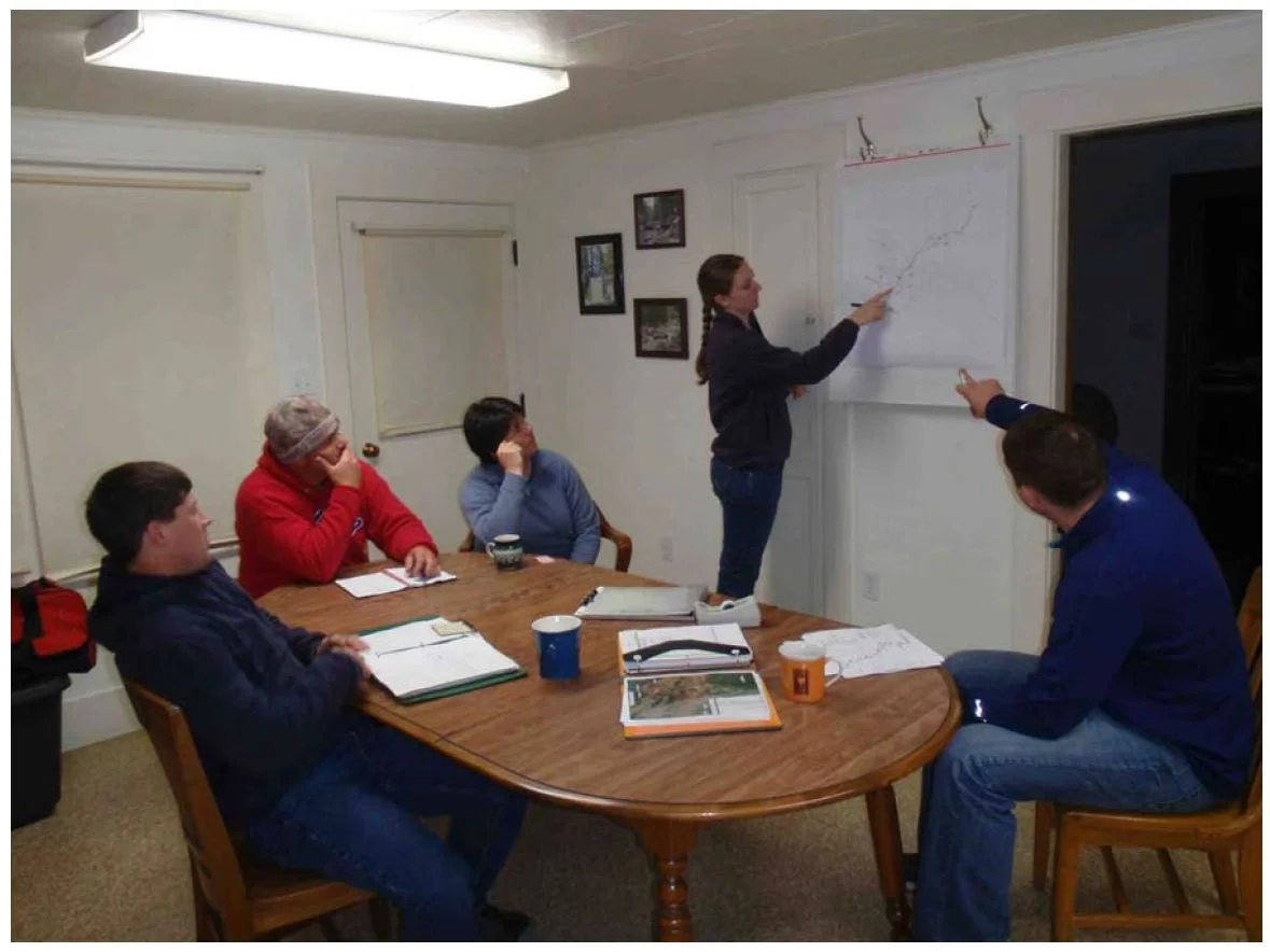 A group of five people sitting around a wooden table in a meeting room, with one woman standing and presenting a graph on a whiteboard, while the others listen and take notes.
