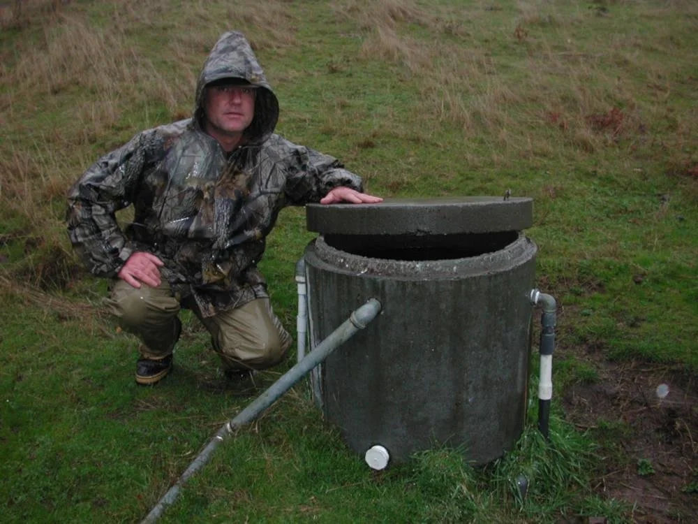 A man in camouflage clothing crouching next to a large, round septic tank with an open lid in a grassy outdoor area.