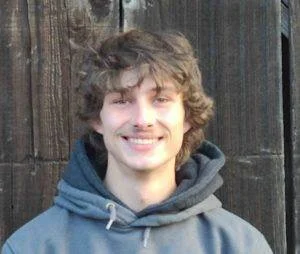 A young man with curly brown hair wearing a gray hoodie, smiling in front of a wooden fence.