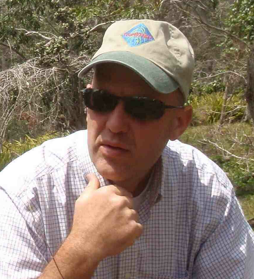 A man wearing a beige hat with a logo, sunglasses, and a checkered shirt in an outdoor setting with bushes and trees.