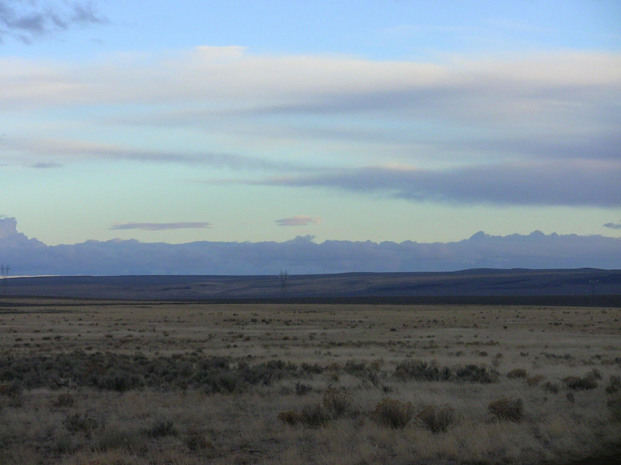 A vast open plain with dry grass and sparse shrubs in the foreground, extending to distant low hills with power lines, under a sky with layered clouds at sunset or sunrise.