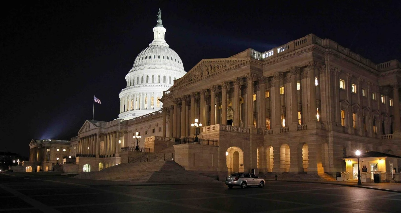 Night view of the U.S. Capitol building in Washington, D.C., illuminated with lights, with an American flag flying nearby.
