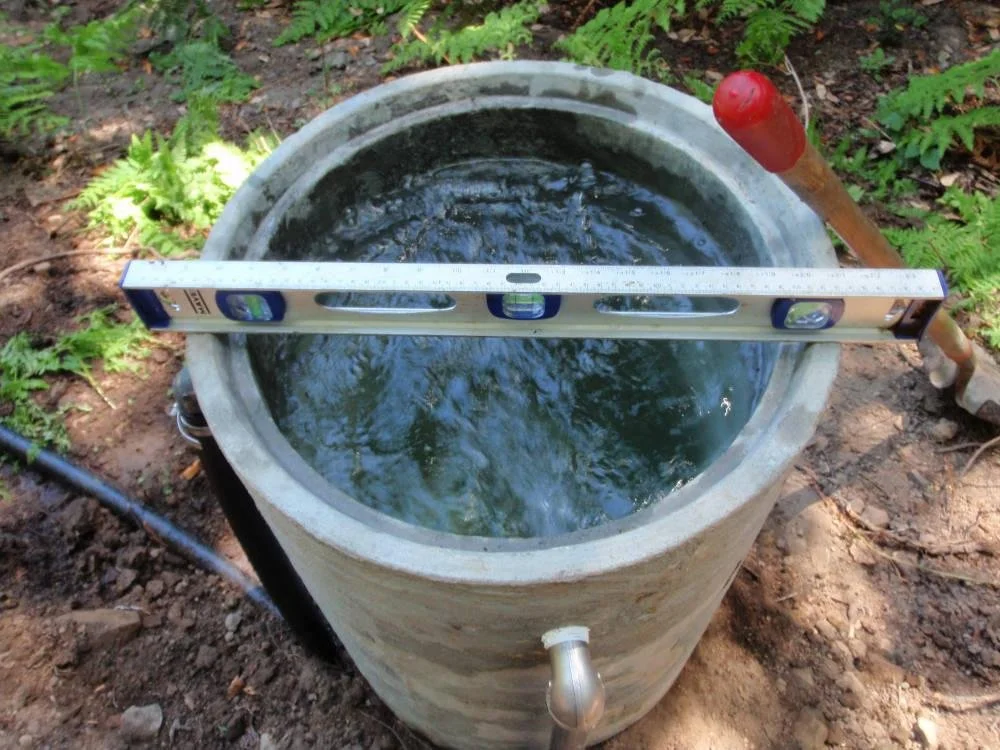A concrete well with water, a level tool on top, and a shovel handle nearby outdoors surrounded by greenery.