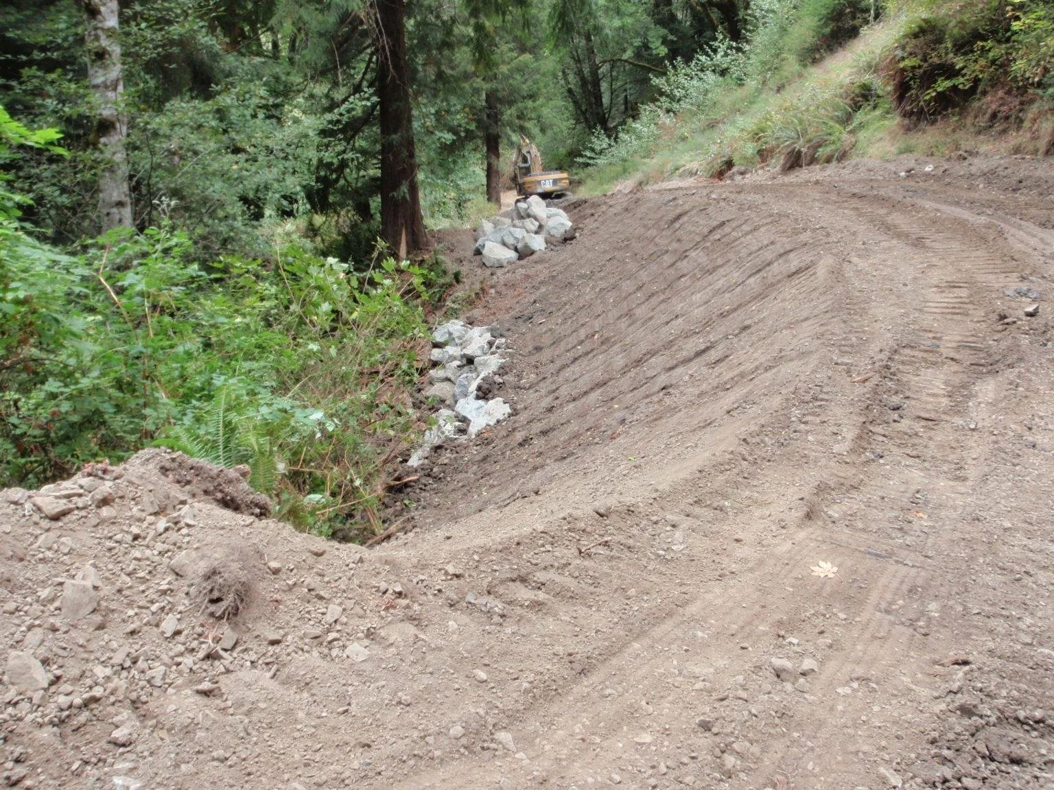 A construction site in a wooded area with a yellow construction vehicle working on a dirt road, which is being prepared or repaired with visible tire tracks and loose dirt.