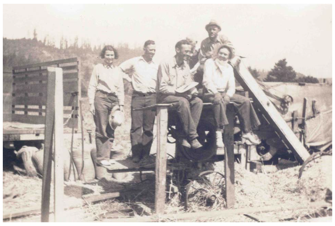 Five people sitting and standing on a farm tractor, with a rural landscape in the background.