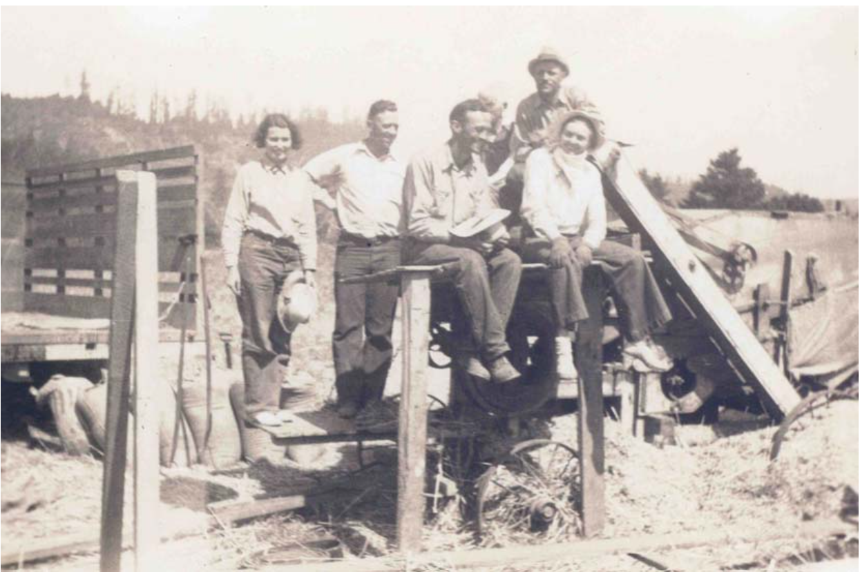 Black and white vintage photograph of six people, three men and three women, sitting and standing on a wooden farm tractor or machinery in a rural outdoor setting.