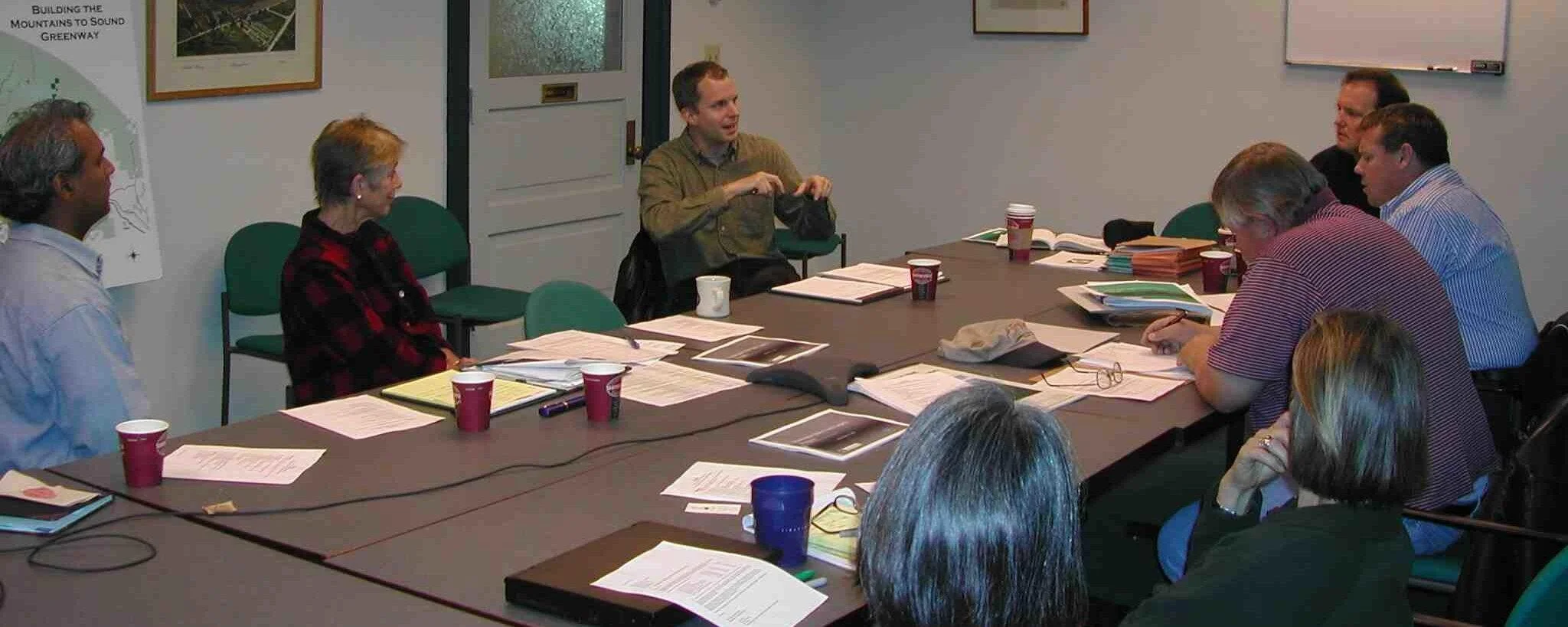 A group of people seated around a conference table engaged in a discussion, with documents, cups, and a telephone on the table.