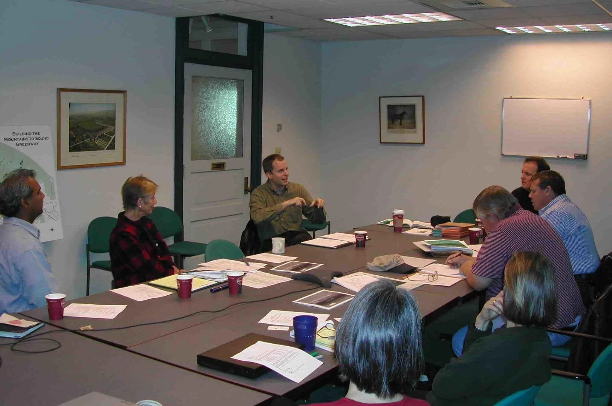 A group of people sitting around a conference table engaged in a meeting, with documents, notebooks, and coffee cups on the table.