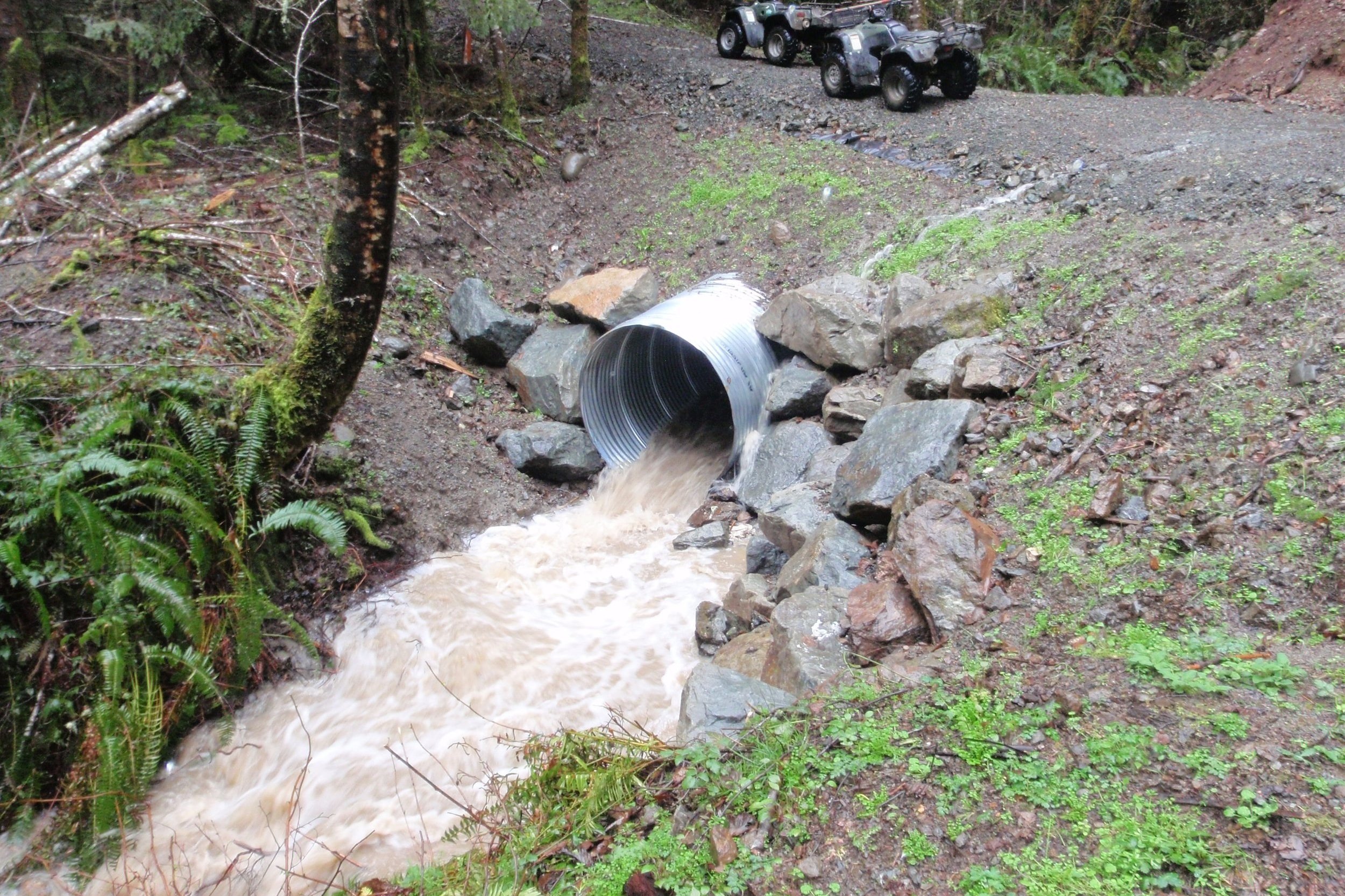 A large metal pipe on a hillside discharges muddy water into a small rocky stream surrounded by green vegetation and trees, with a dirt road and several all-terrain vehicles parked nearby.