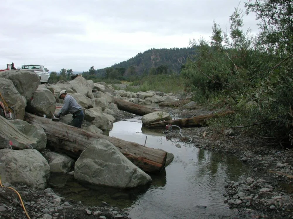 A man working near a small creek with large rocks and logs, with cars parked on a rocky area in the background, and hills and cloudy sky in the distance.