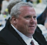 Portrait of a middle-aged man with gray hair, wearing a dark suit and white shirt, sitting outdoors at a formal event.