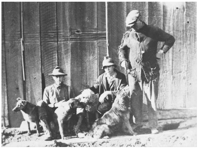 Three men with dogs in front of a wooden fence or barn wall. The men are wearing hats and casual outdoor clothing, and two are sitting while one stands.