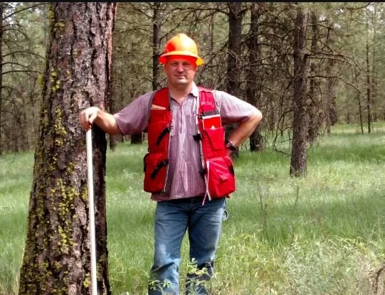 Man in a red hard hat and red vest standing in a forest with trees and grass, holding a measuring stick.