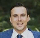 Professional headshot of a smiling man in business attire outdoors with greenery in the background.