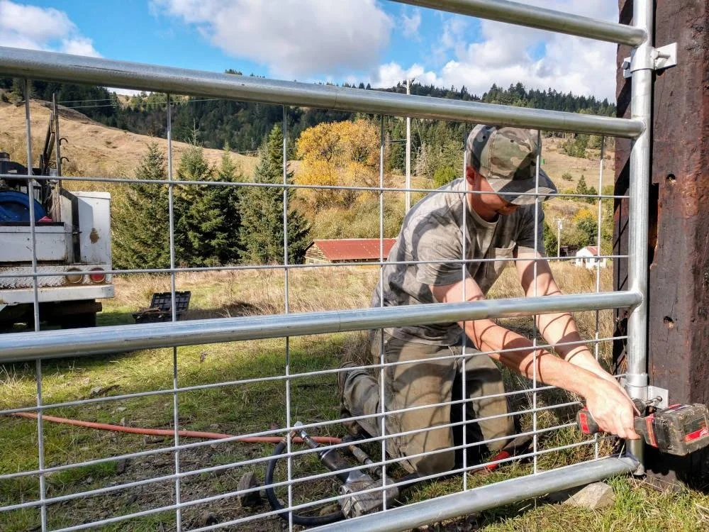 A man working on a metal gate outdoors, kneeling on grass with a power drill in hand, wearing a camouflage cap, sunglasses, and casual clothes. There is a truck and trees in the background with a hill and clear sky.