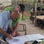 Two men are outdoors looking at papers on a table. One man is writing or pointing at the papers.