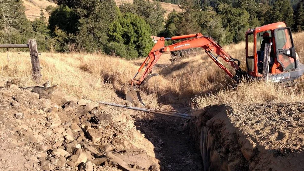 A small orange excavator digging a trench through dirt and rocks in a grassy field with trees in the background.