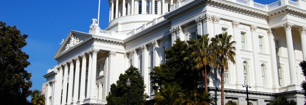 White neoclassical government building with columns and decorative statues, trees in front, with a clear blue sky background.
