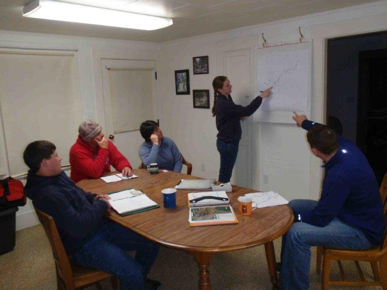 A woman is giving a presentation with a graph on a whiteboard, while four people, sitting at a table with notebooks and mugs, watch and listen.