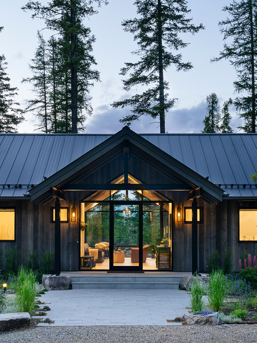 Modern house with a glass entrance door, surrounded by greenery, with tall trees in the background and warm interior lighting visible through the windows.