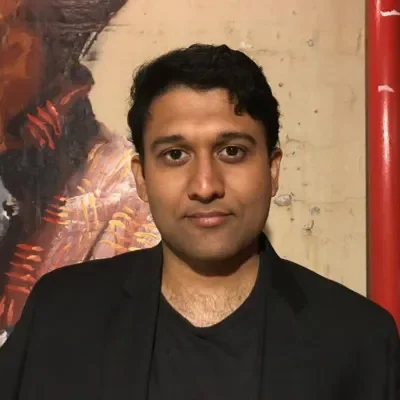 A man with dark curly hair, dressed in a black blazer and shirt, standing in front of a painted wall with a low intensity smile.