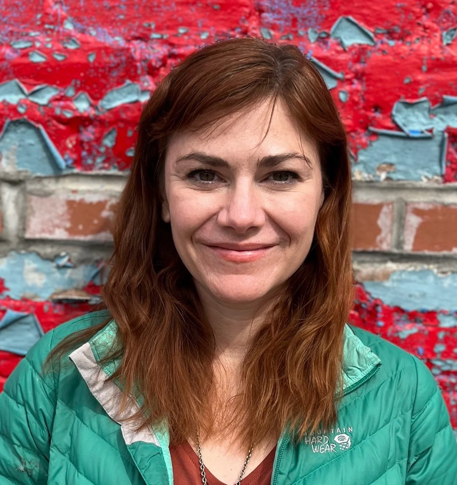 A woman with long red hair smiling in front of a colorful mural with red and blue textures and patterns.