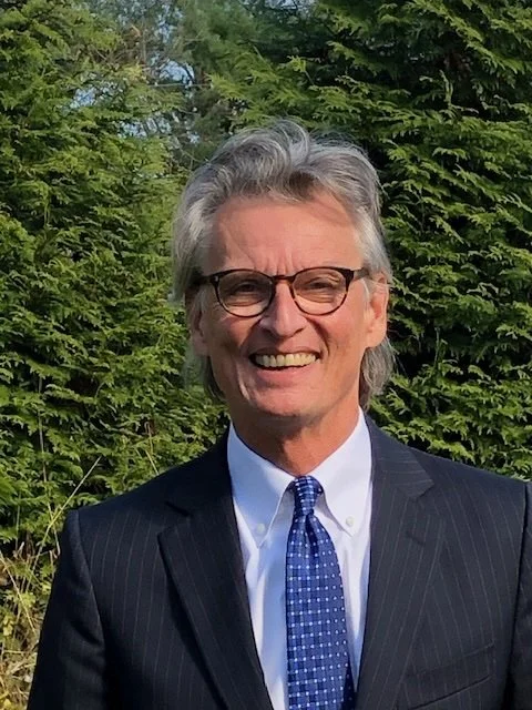 A smiling man with gray hair, glasses, wearing a dark pinstripe suit, white shirt, and blue tie, standing outdoors with green trees in the background.