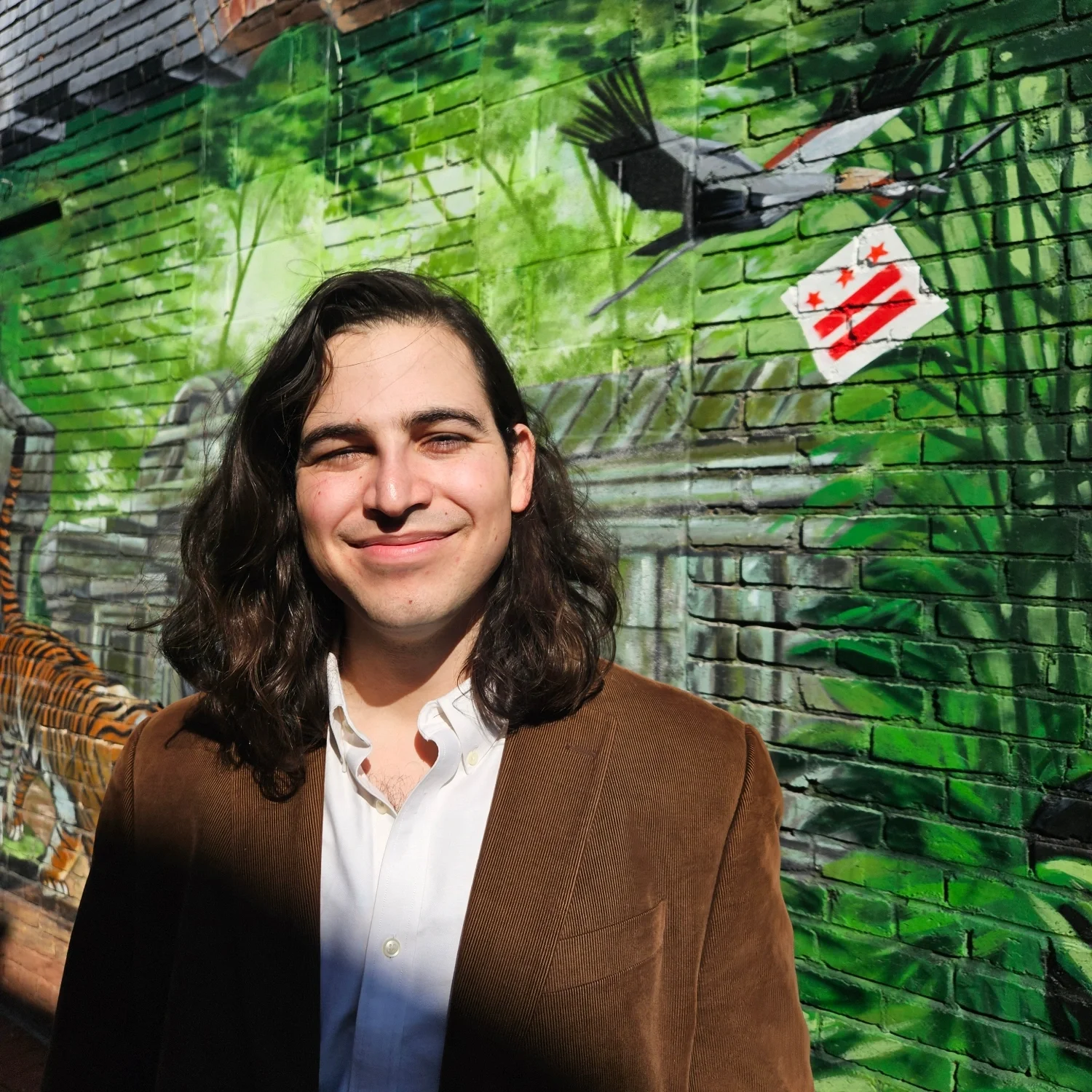 Young man with long dark hair smiling outdoors in front of a colorful mural painted on a brick wall, featuring green foliage, a bird, and a red and white sticker or flag.