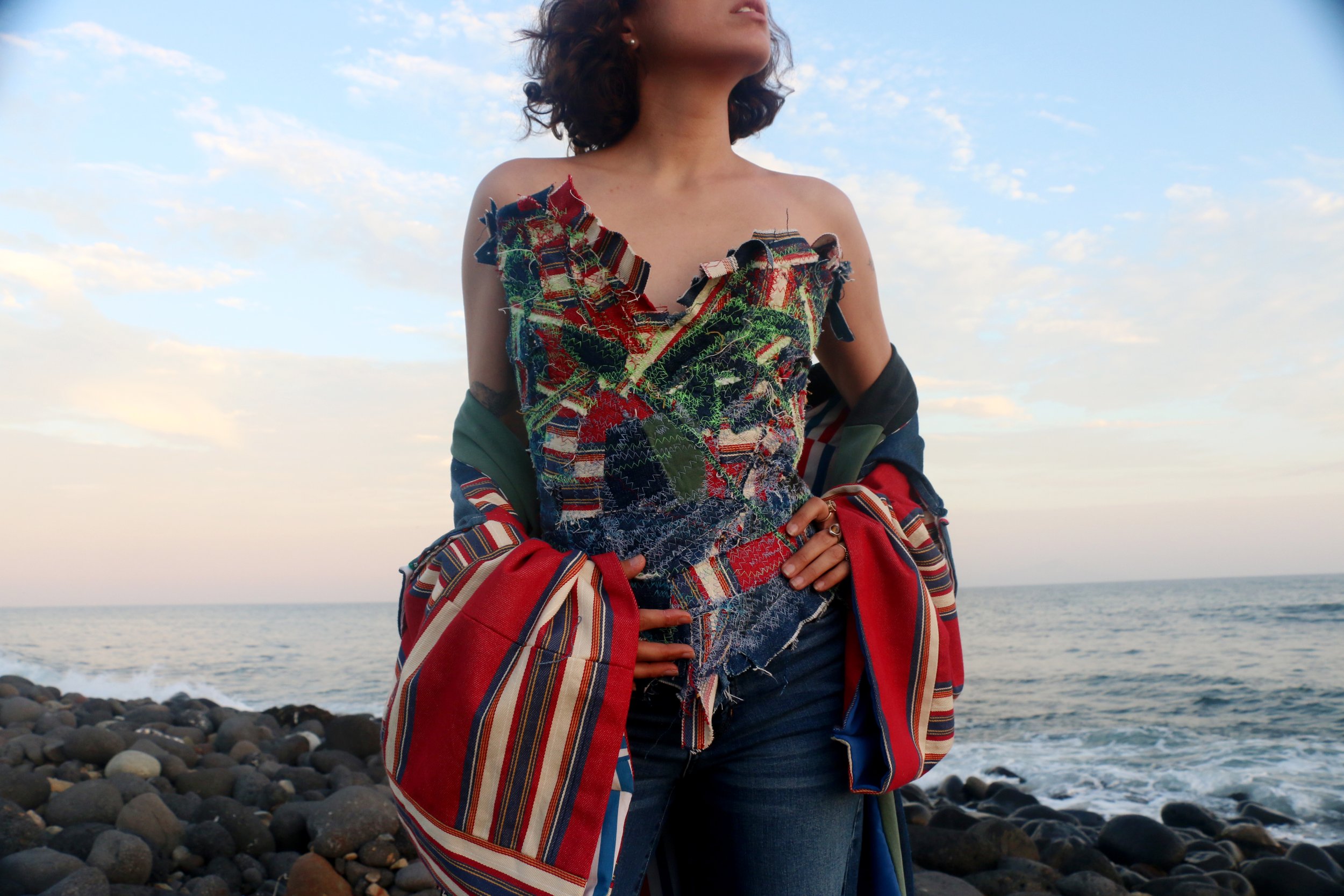 A model wearing a sculptural corset assembled from multicolored denim scraps and exposed stitching beside the ocean.
