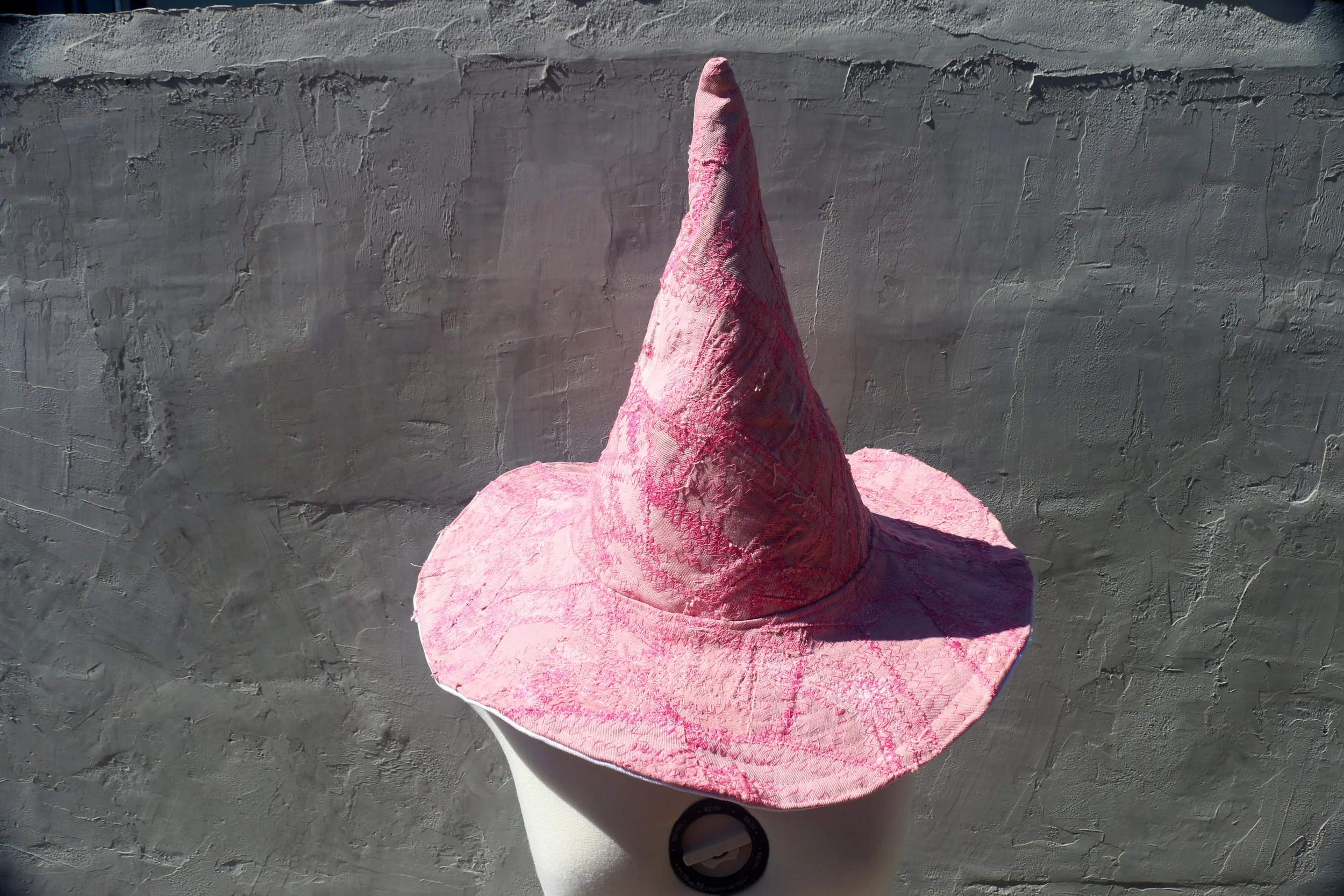 Sculptural pink patchwork denim witch hat with a tall pointed crown and wide brim, displayed against a textured gray wall.