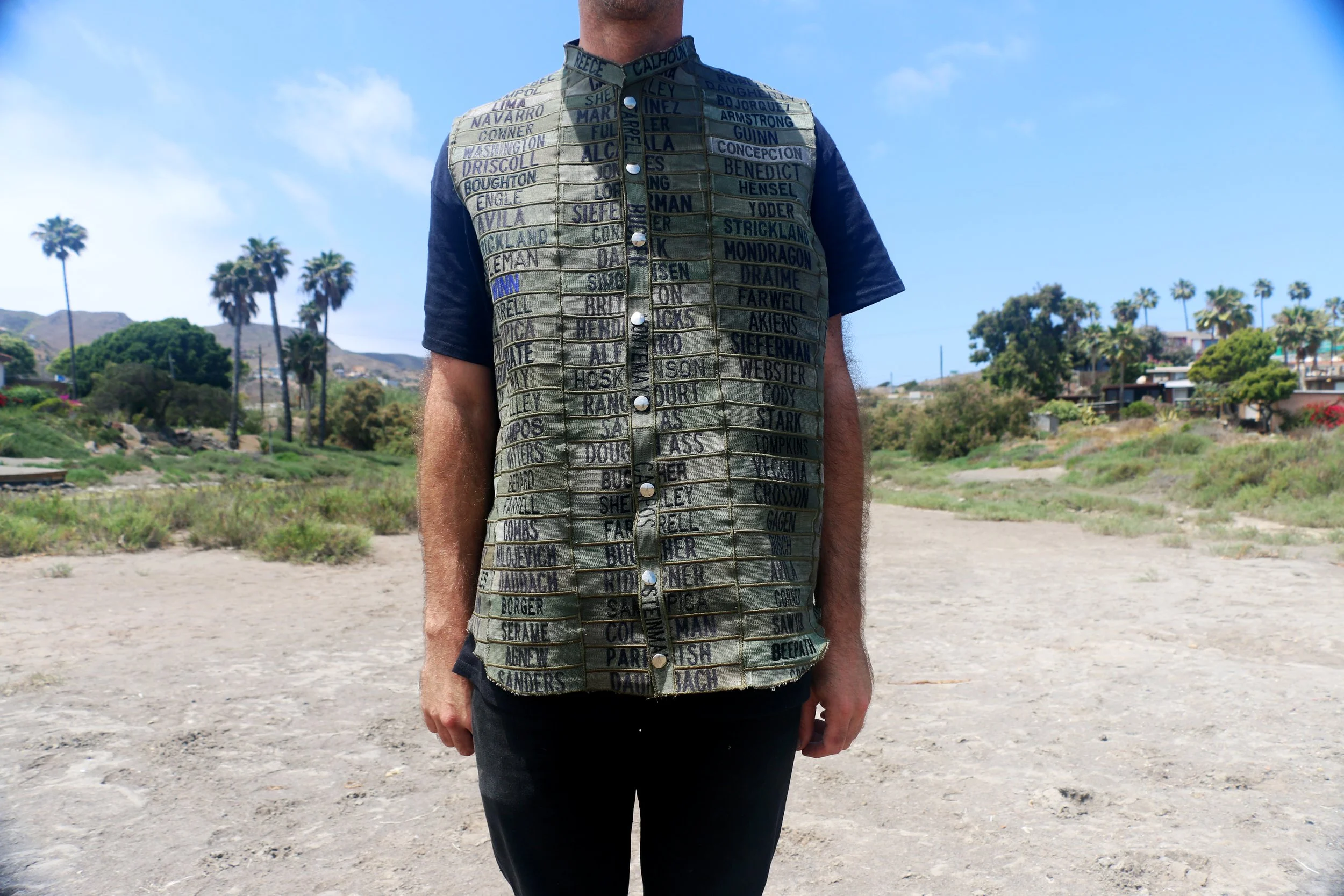 Front view of a model wearing an olive vest constructed from rows of embroidered U.S. Navy name patches and metal snap closures.