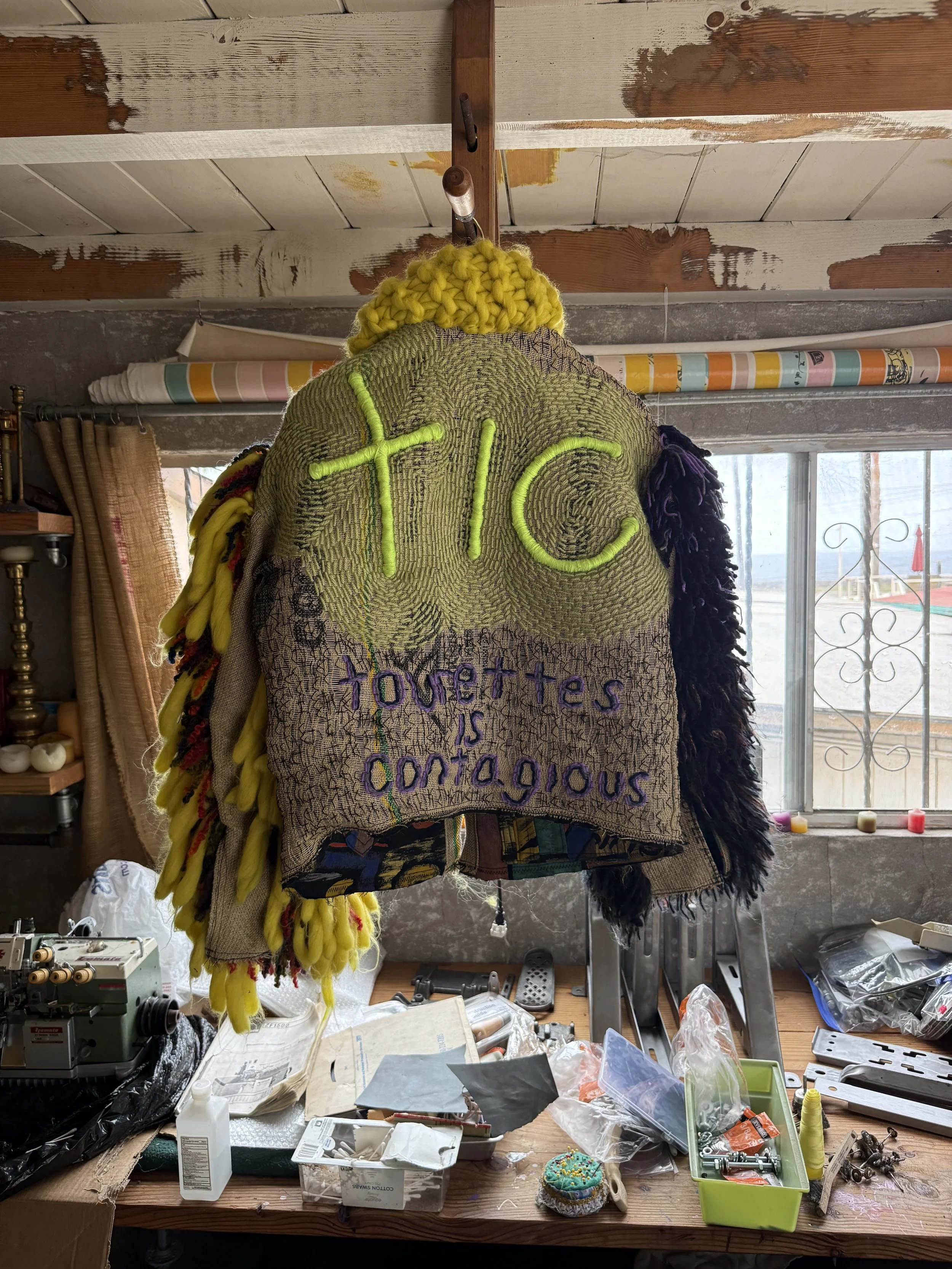 Back view of a sculptural black and yellow TIC jacket with a reclaimed-textile panel embroidered “TIC” and “Tourette’s is contagious.”