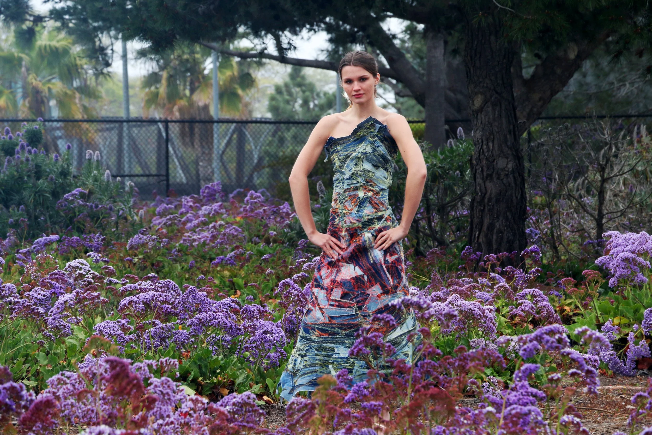 A model wearing a sculptural patchwork denim corset cocktail dress among purple flowers, showing its fitted bodice and full-length silhouette.