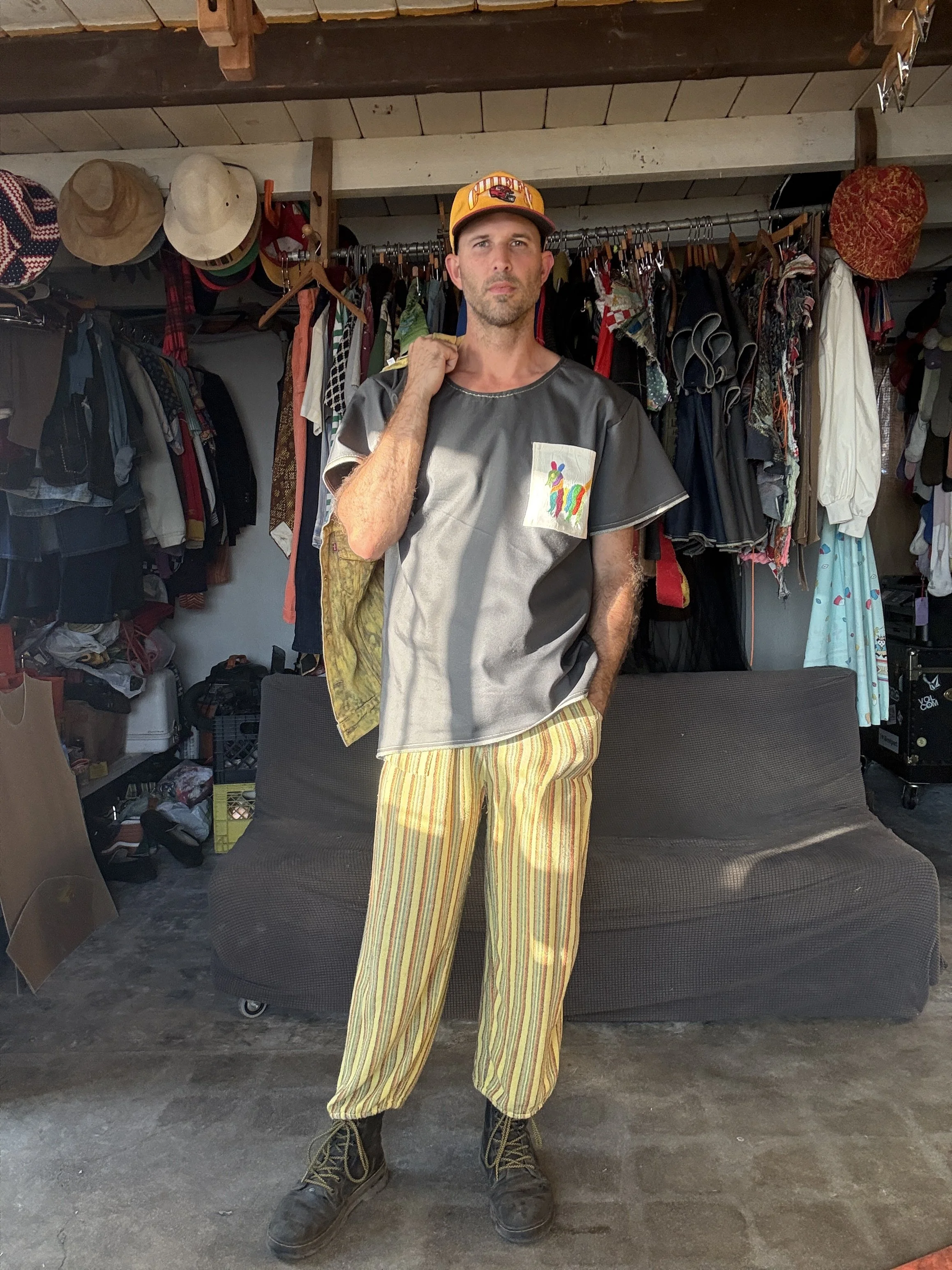 Addison Stonestreet wearing a charcoal denim T-shirt with a colorful embroidered pocket and striped trousers inside a clothing-filled studio.