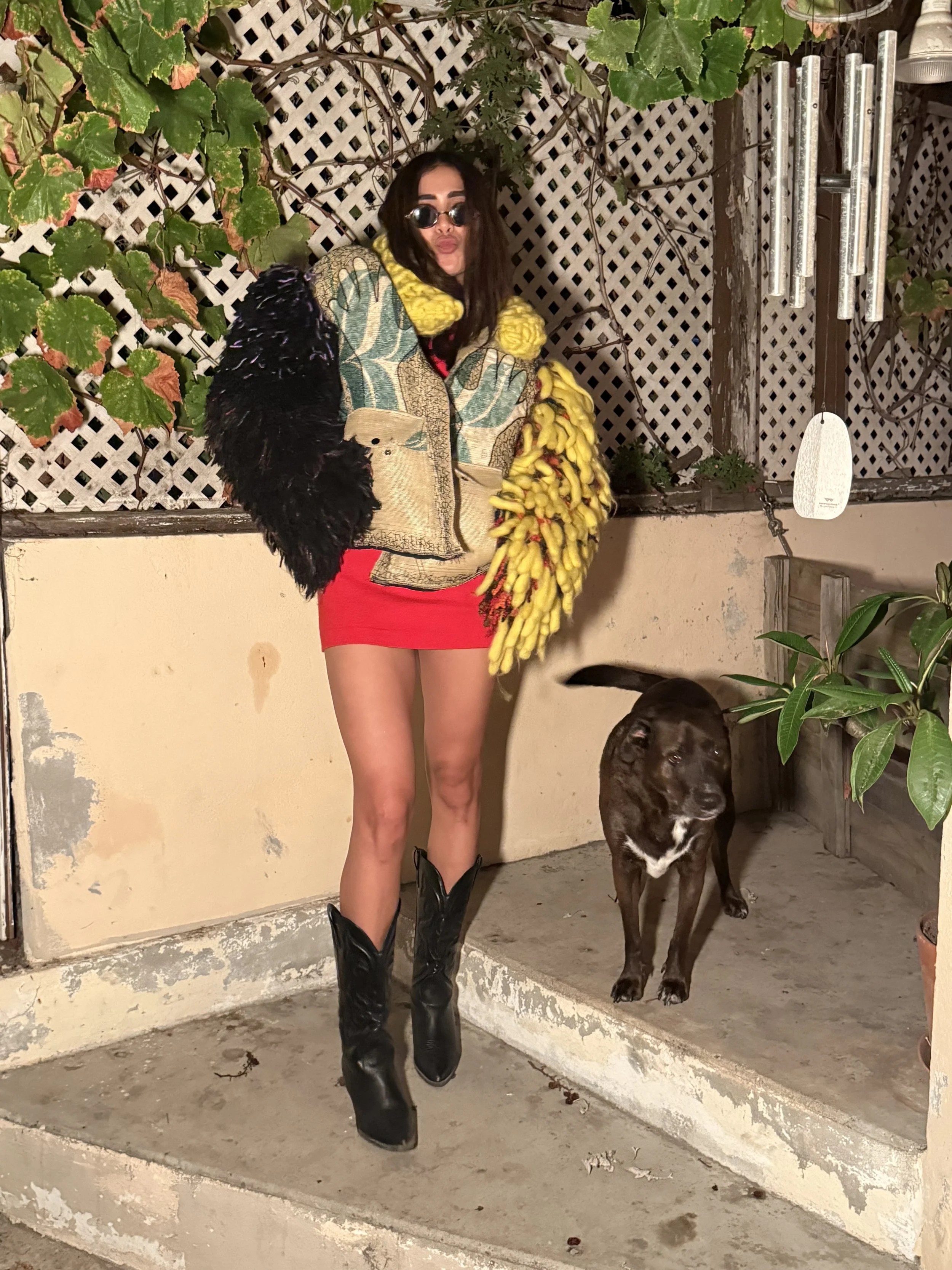 A model wearing a sculptural patchwork TIC jacket with black and yellow shaggy sleeves, red shorts, and black boots outdoors beside my dog.