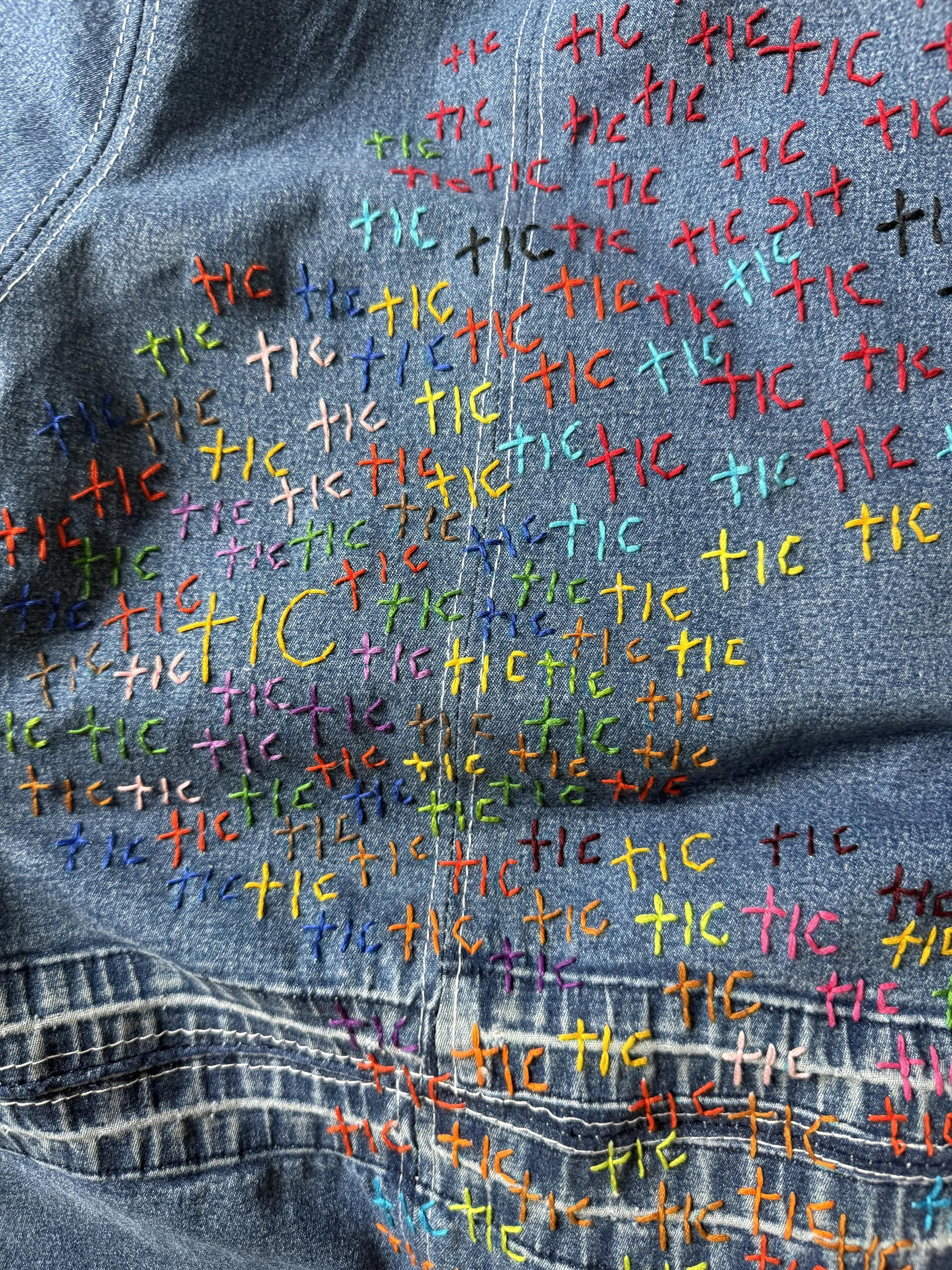 Close-up of blue denim densely embroidered with repeated multicolored TIC lettering across the fabric and pocket.