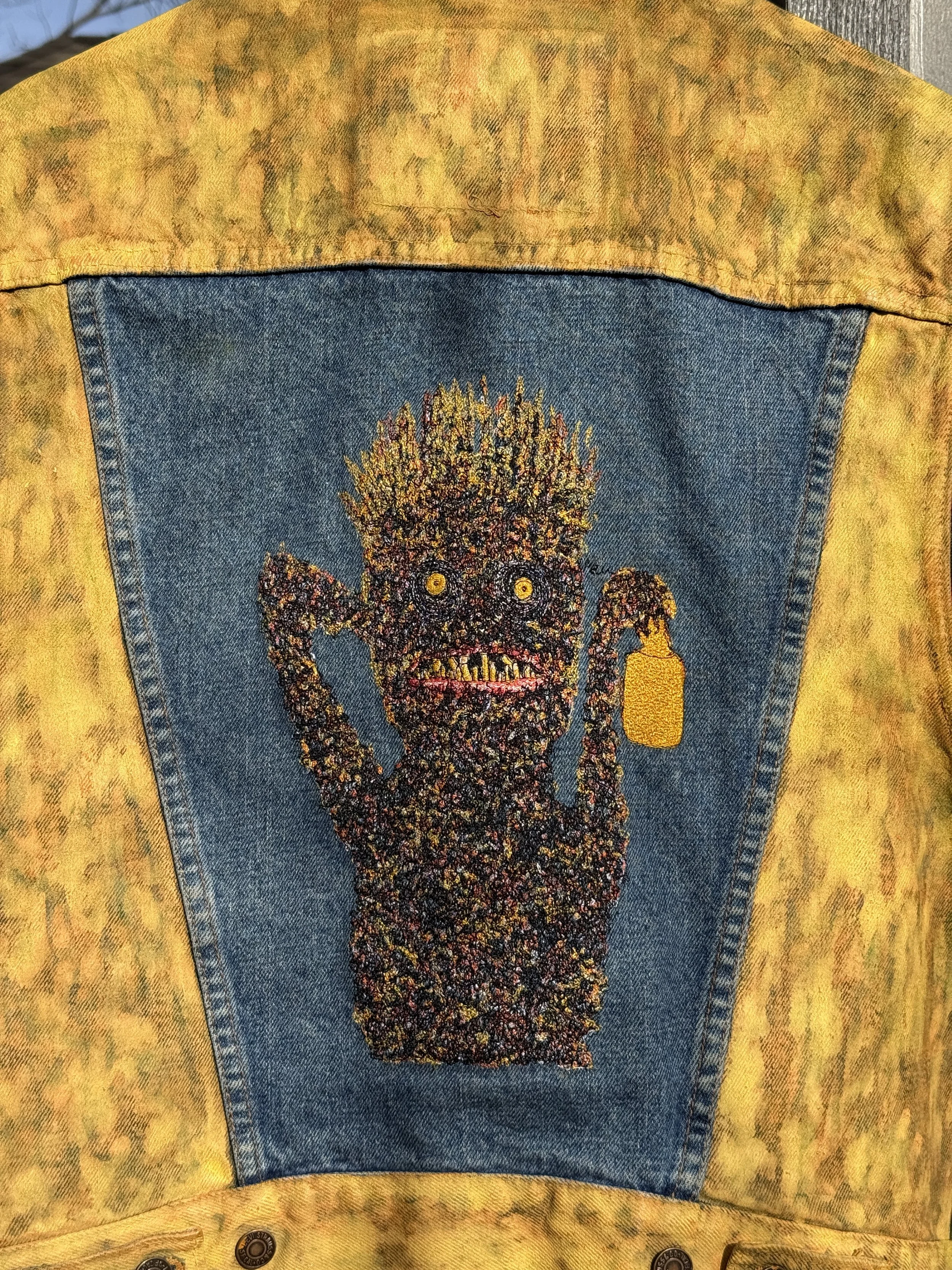 Back view of a yellow sleeveless denim vest with a blue denim inset and embroidered creature artwork.