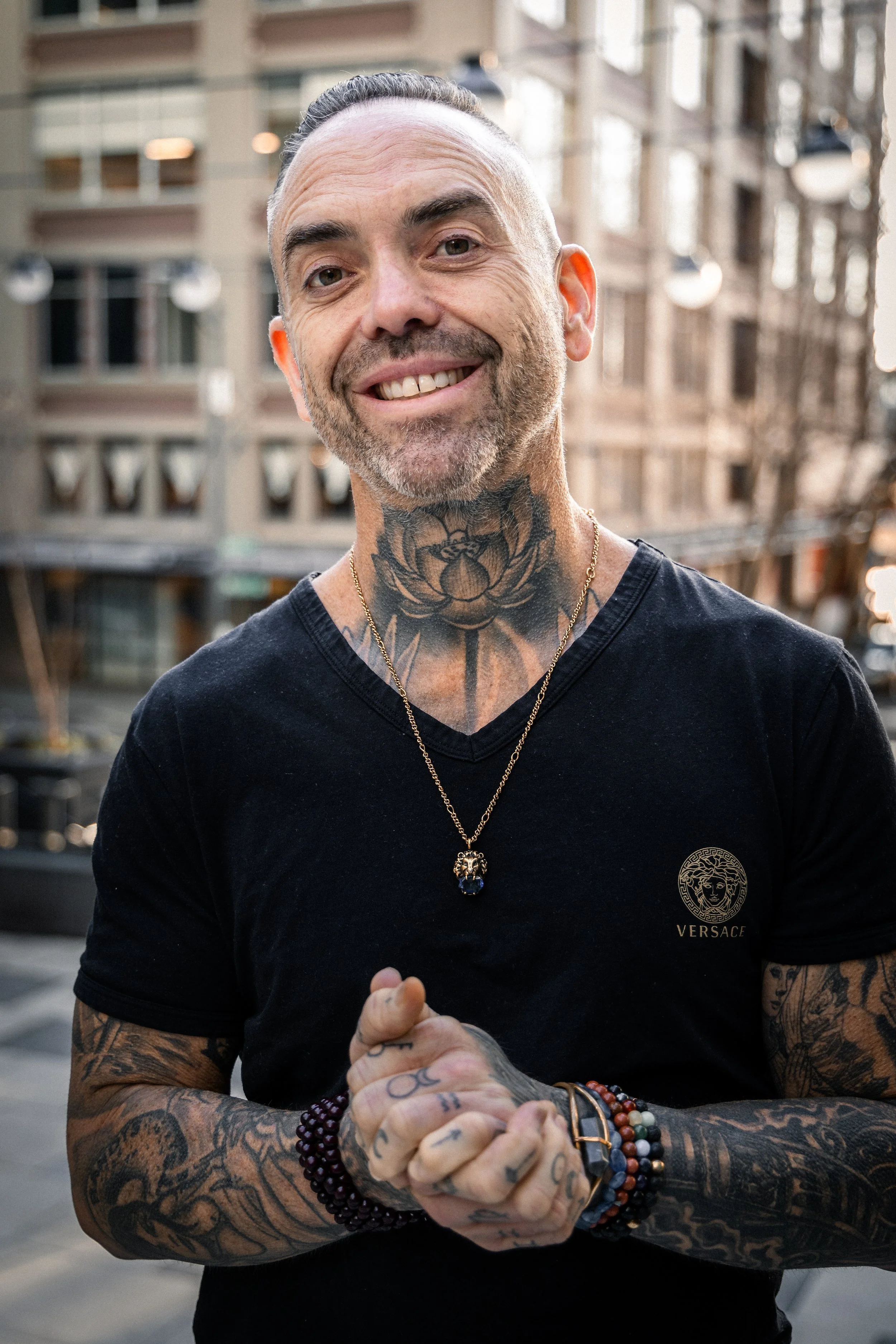 A smiling man with tattoos, wearing a black Versace t-shirt, a gold necklace, and bracelets, standing outdoors in an urban setting.
