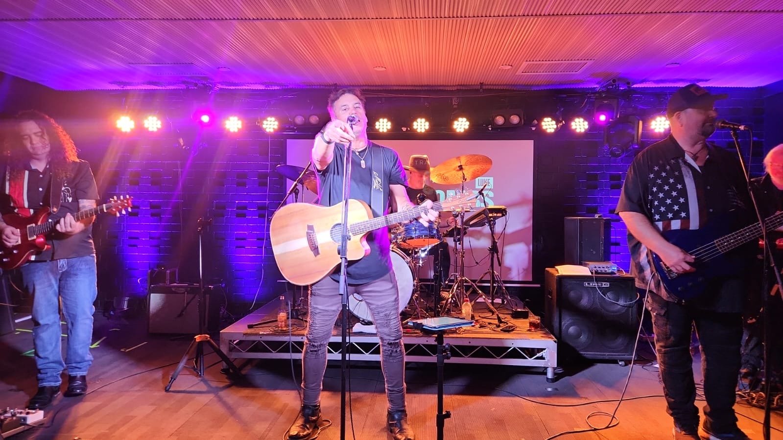 Band performing on stage with guitarist, drummer, and singer playing acoustic guitar under colorful lights.