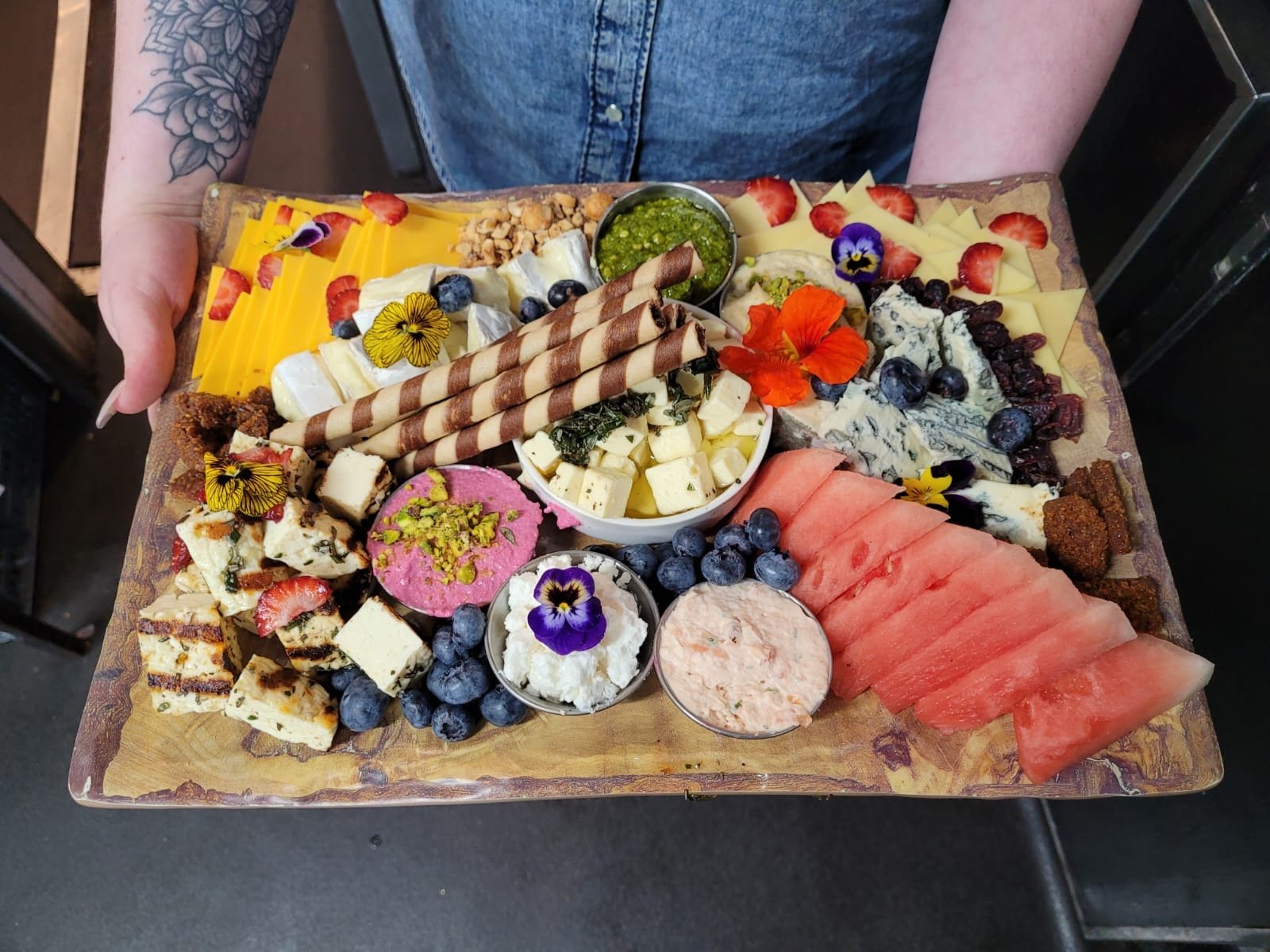 A charcuterie board with assorted cheeses, fruits, crackers, desserts, and spreads. Includes cheese cubes, sliced watermelon, blueberries, strawberries, breadsticks, edible flowers, and various dips in small bowls. Person holding the board visible.