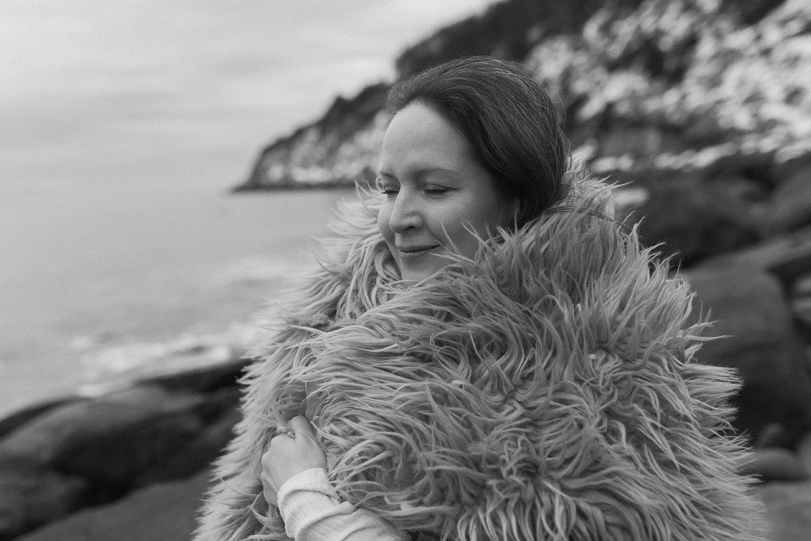 Black and white photo of a woman wrapped in a furry coat, standing near a rocky coastline.