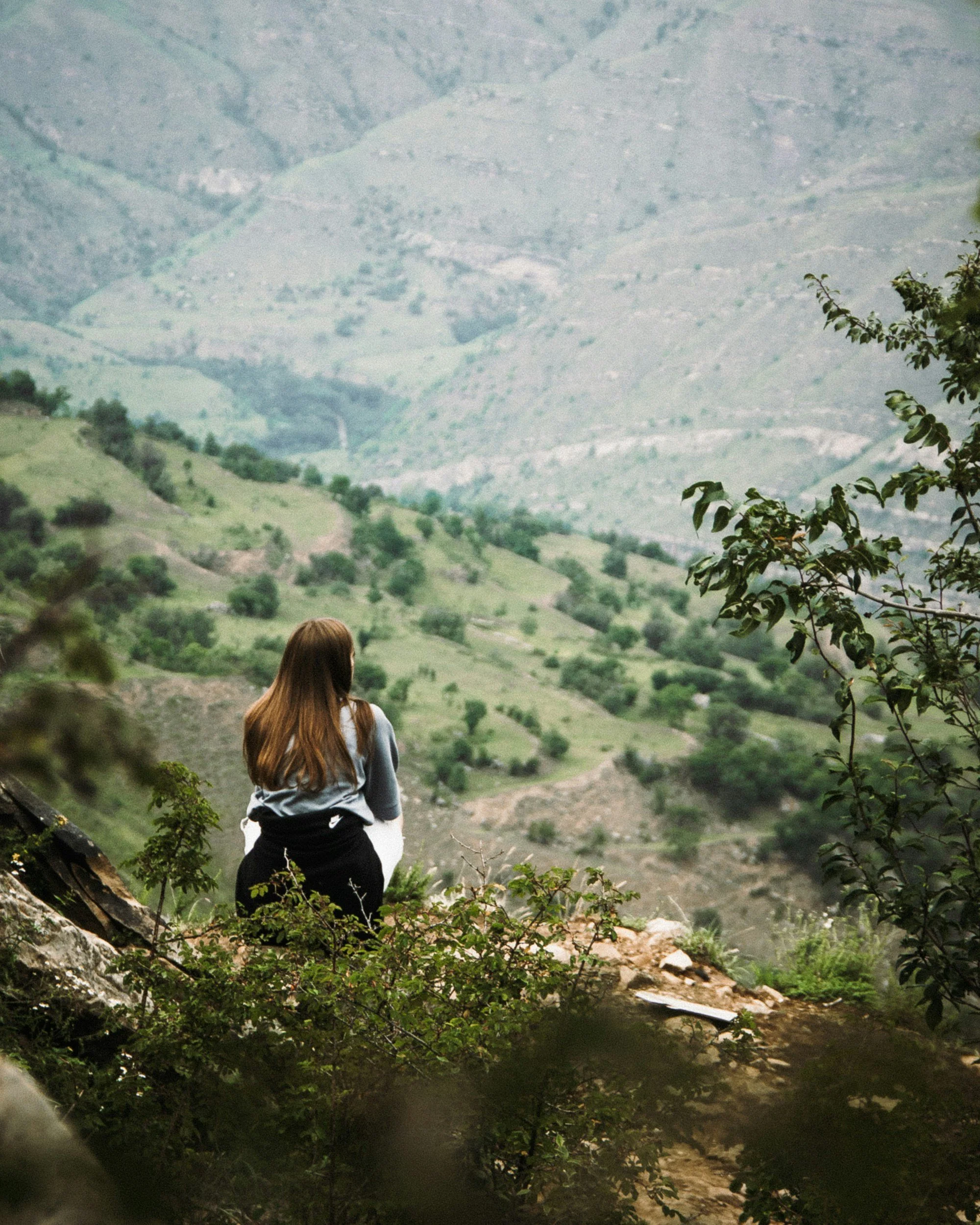 Woman sitting on a rocky ledge overlooking green, hilly landscape with trees and mountains in the distance.