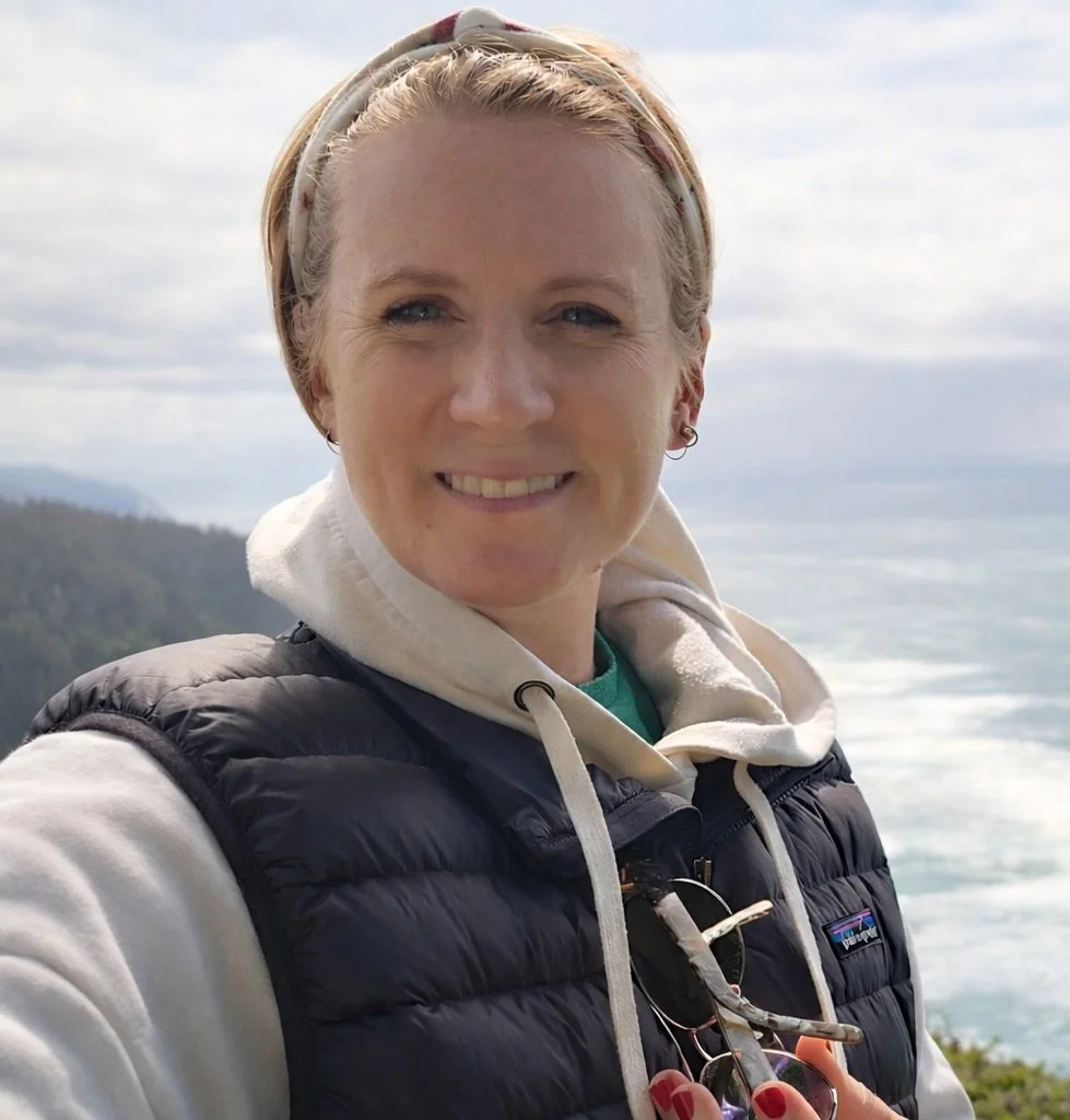 A woman with short blond hair, light-colored eyes, and light skin, smiling outdoors near a coastal area with trees and water in the background. She is wearing a beige hoodie, a black puffer vest, and a headband.